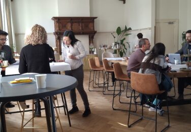 several folks in a meeting room, sitting around tables and drawing on paper or chatting with eachother