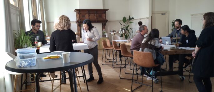 several folks in a meeting room, sitting around tables and drawing on paper or chatting with eachother