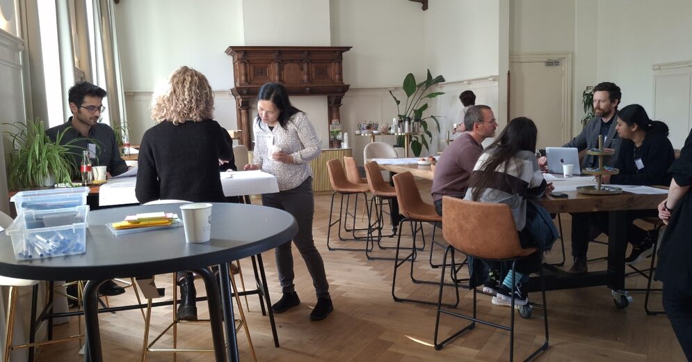 several folks in a meeting room, sitting around tables and drawing on paper or chatting with eachother